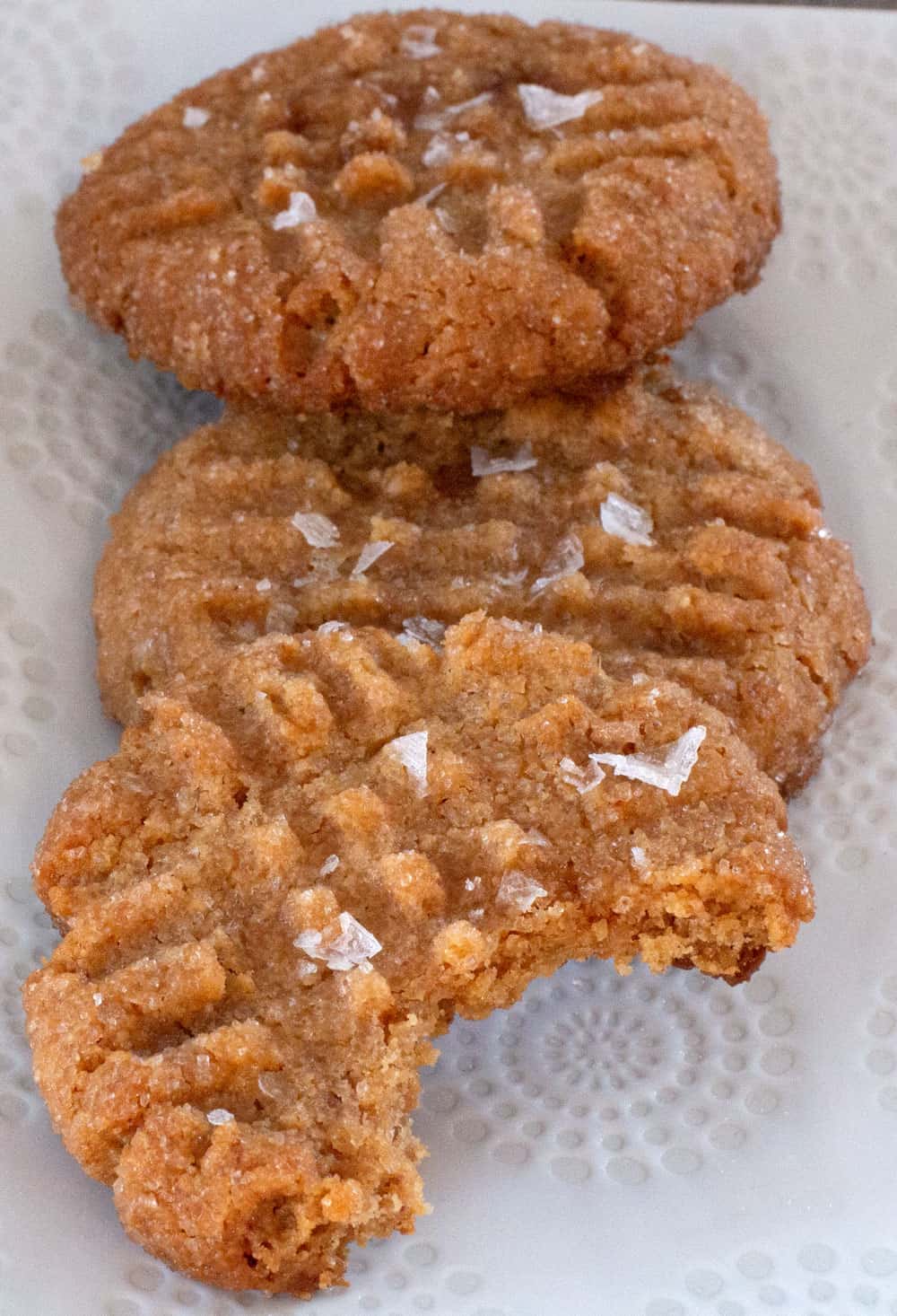 3 vegan peanut butter cookies topped with flaked salt, with bite taken out of one in front